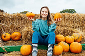 Smiling young woman sitting on straw bales and holding pumpkins. selecting best one for Thanksgiving and Halloween holidays decoration on agriculture farm. Pumpkin harvest. Autumn fall festive mood