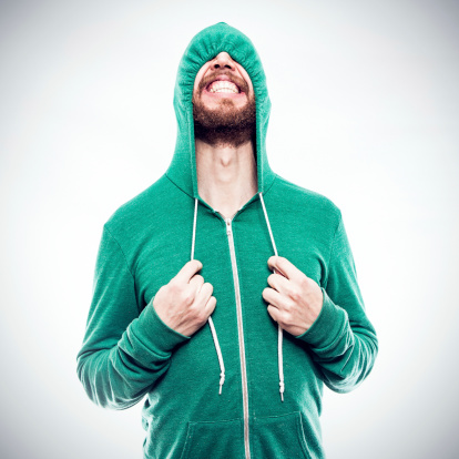 A bearded hipster young man pulls the drawstrings on his green hoodie, partially concealing the funny face he is making, complete with a cheesy grin.  Square crop.