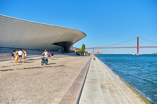 Lisbon, Portugal - August 08, 2022: MAAT - Museum of Art, Architecture and Technology is a modern building on a bank of Tagus river with 25th of April bridge in background. Lisbon, Portugal