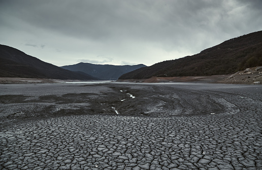 Photo captures the drying mud and cracks in a river valley under cloudy weather, affected by a hydroelectric power plant.