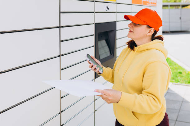istock Young woman in red hat picks up parcel from automatic post office machine, Courier standing with phone and small box. Concept of fast delivery to automatic self lockers 1619302749