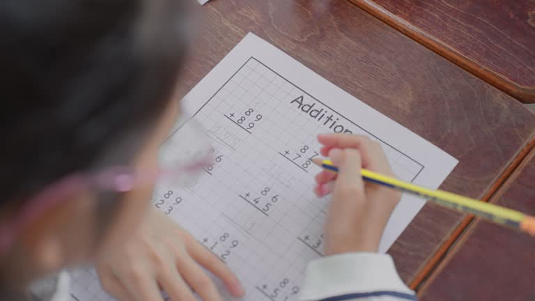 Concentrated elementary school kid wearing eyeglasses completing number addition lesson in mathematics class at school, student learning number addition on paper, self-learning