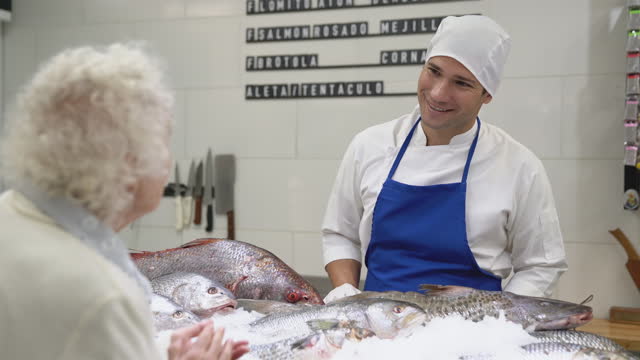Young fishmonger helping senior female customer choose fresh seafood products from the cabinet display
