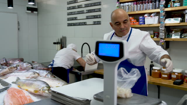 Mature fishmonger adding calamari to a plastic bag for a customer while unrecognizable employee works at background