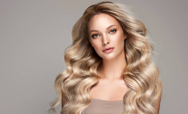 Young, blond haired beautiful model with long, curly, well groomed hair. Hair care and hairdressing art. Portrait of beautifully looking young blond haired woman dressed in a delicate evening makeup.Perfect model straightly gazing by kind tender look at viewer. Beauty elegance hairstyling. blond hair stock pictures, royalty-free photos & images