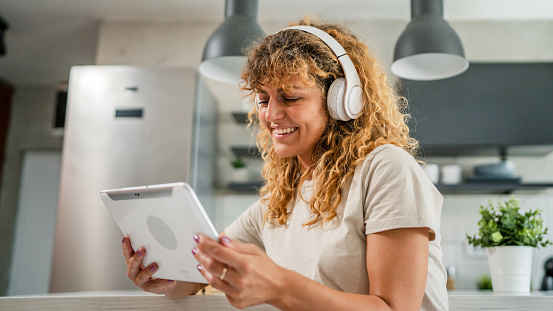 One caucasian woman with curly hair sit at home use digital tablet and headphones to watch movie or series online stream or to have video call happy smile have a good time copy space