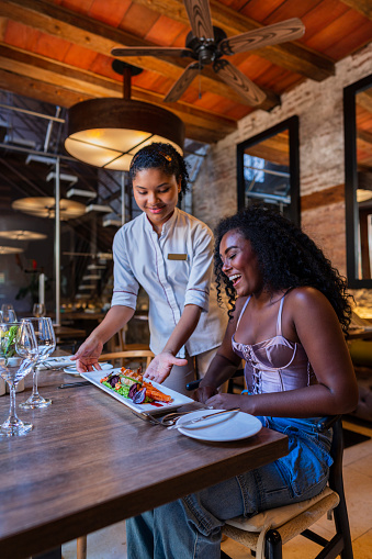 Afro-haired Latina woman, a waitress by profession, is in the restaurant she works for, dressed in a uniform, serving an Afro-Latin woman who goes to the restaurant to try delicious dishes