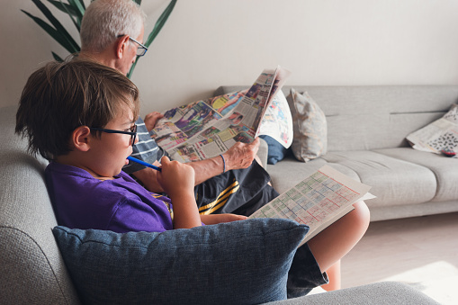 A boy is solving sudoku while his grandfather is reading the newspaper next to him. Grandfather and grandchild are relaxing in their home.