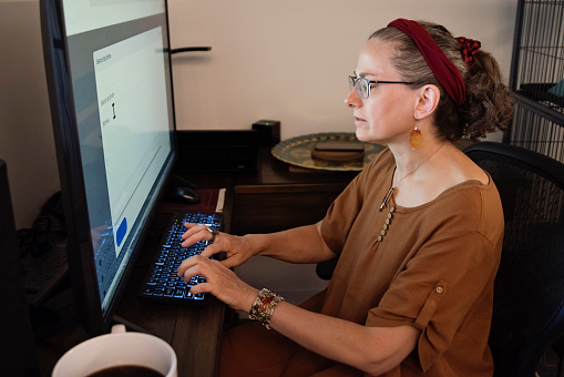 Woman living with a visual disability working at home. She use a tv screen for her computer, to have larger letters to see. Big coffee cup in the foreground. She is in her early fifties, and is wearing casual clothes. Horizontal waist up indoors shot with copy space. This was taken in Quebec, Canada.