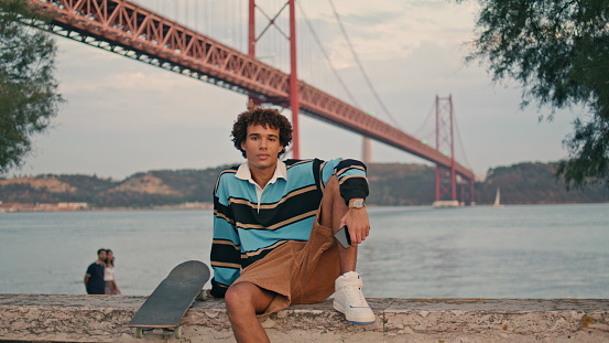 Young man sitting skateboard at water view coast portrait. Thoughtful skater guy looking camera at Tagus river evening background closeup. Sporty hipster thinking at famous Lisbon place zoom out