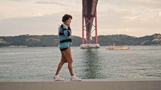 Relaxed tourist speaking smartphone at Lisbon embankment. Curly guy talking mobile at 25th of April bridge background. Contemporary teenager walking at river view outdoor. Urban lifestyle concept