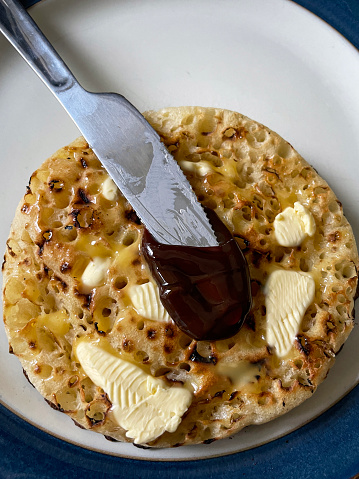 Stock photo showing close-up, elevated view of a blue rimmed plate containing freshly toasted, warm, buttered crumpet with melting butter.