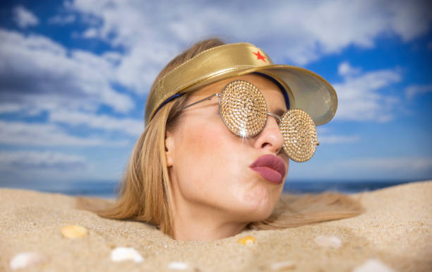 A close up of a young woman’s face. Her hair is wind blown from salty water. She is wearing reflective sun glasses that show she is looking at palm trees