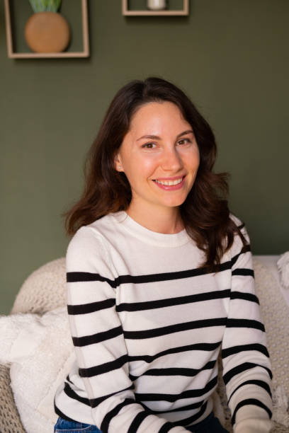 Young attractive caucasian woman sits alone on the sofa in the living room during the day and smiles cutely while looking at the camera Young attractive caucasian woman sits alone on the sofa in the living room during the day and smiles cutely while looking at the camera. High quality photo cutely stock pictures, royalty-free photos & images