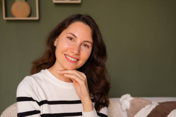 Closeup portrait of a young attractive caucasian woman sitting alone on the sofa in the living room during the day and smiling cutely looking at the camera Closeup portrait of a young attractive caucasian woman sitting alone on the sofa in the living room during the day and smiling cutely looking at the camera. High quality photo cutely stock pictures, royalty-free photos & images