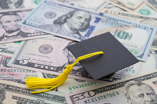 A young female graduate turns around as she poses for a portrait.  She is wearing a gown and cap and holding her diploma as she stands with her peers who have their backs to the camera.