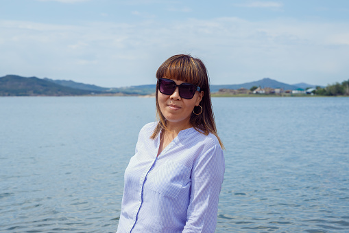 young asian kazakh bodypositive female woman in sunglasses, blue shirt on lake beach, mountains. front view portrait