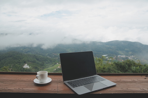 Computer Monitor, Keyboard, coffee cup and Mouse with Blank is on the work table at the sky mountain river and trees front view background.