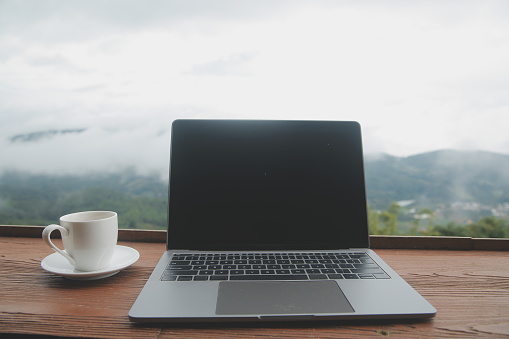 Computer Monitor, Keyboard, coffee cup and Mouse with Blank is on the work table at the sky mountain river and trees front view background.