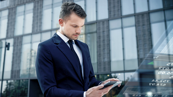 Businessman using stock buying app on his digital tablet while standing in front of modern office building.