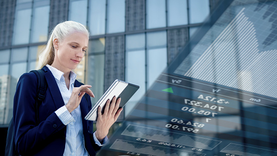 Young businesswoman using trading app on her digital tablet while standing outside modern office building.