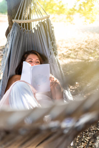 Woman enjoying a book in a hammock on a sunny day, with straw hat and garden background.