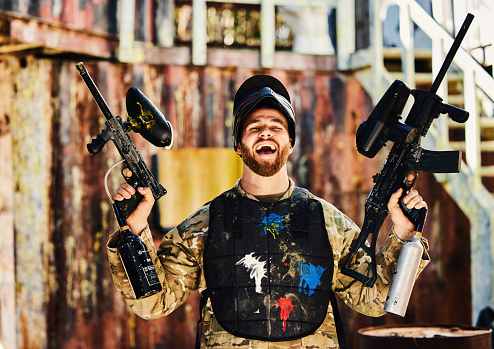 Paintball, laughing or portrait of happy man with guns in shooting game playing or celebrates battlefield mission. Crazy or funny soldier with army weapons gear winning military challenge competition