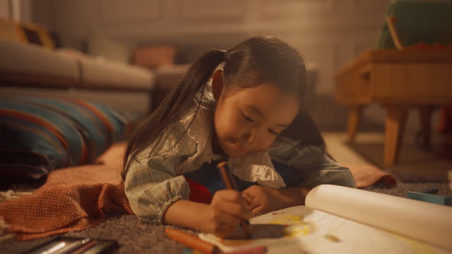 Evening Portrait of Cute Little Girl Drawing while Lying on the Floor in Living Room. Talented Korean Child Being Creative, Coloring Picture, Preparing a Become Famous Artist. Static Shot