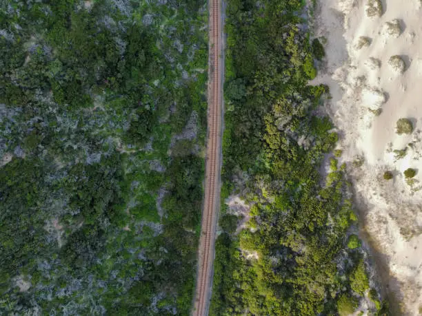 Aerial shot of a coastal railroad cutting through lush green bushes and plants next to a white pristine beach with dunes with some green grass growing out of the dunes.