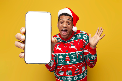 african american guy in christmas clothes and santa hat shows blank smartphone screen on yellow isolated background, man advertises phone display in christmas sweater and is amazed