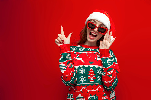 cute girl in christmas sweater and santa hat in glasses shows her hand to the side on red background, shocked woman in christmas clothes advertises copy space