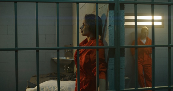 One female prisoner in orange uniform stands behind metal bars, another sits on the bed in prison cell. Women serve imprisonment terms for crimes in jail. Depressed inmates in detention center.