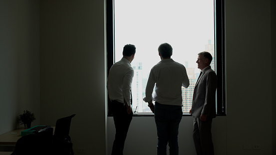 Group of businessmen talking plan together standing beside windows at office. Silhouette colleague businessman discussion idea plan work at workplace. Collaboration teamwork partnership organization.