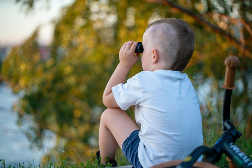 Cute boy looking through binoculars while sitting on the grass, rear view