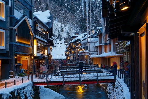 Night scenery of Ginzan Onsen , a famous hot spring town and tourist destination in Obanazawa, Yamagata, Japan, with bridges over a stream flanked by old buildings on a cold snowy winter evening

The photograph captures the mesmerizing night scenery of Ginzan Onsen, a renowned hot spring town and popular tourist destination located in Japan. The town is nestled within a picturesque valley surrounded by lush mountains and features traditional wooden ryokan-style buildings lining both sides of a charming stone-paved street. Soft, warm light emanates from the traditional lanterns and the illuminated interiors of the ryokans, creating a cozy and inviting ambiance.

In the foreground of the image, a serene river meanders through the town, reflecting the illuminated structures and adding a sense of tranquility to the scene. The combination of the vibrant reflections and the dark, starry sky overhead creates a captivating contrast that highlights the beauty of the town's architecture and natural surroundings.

Ginzan Onsen is famous for its historic and preserved atmosphere, evoking a sense of nostalgia for a bygone era. The photograph perfectly captures this timeless charm, inviting viewers to immerse themselves in the enchanting atmosphere of this charming hot spring town, making it a must-visit destination for travelers seeking a blend of cultural authenticity and relaxation.