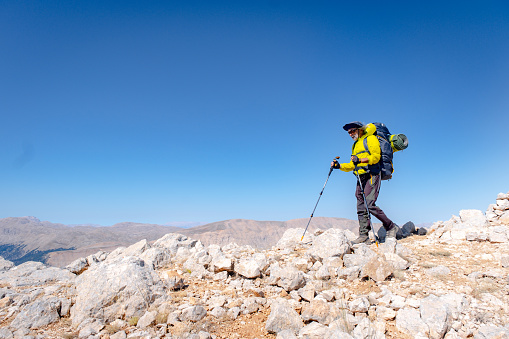 White long-haired man, aged 60-65. He is vigorous, hiking, active lifestyle with backpack, walking pole. He has a yellow windbreaker on him. He is hiking in a mountainous region at high altitude. Mediterranean region. At the top of the mountain. Model looks full length. Light blue sky.