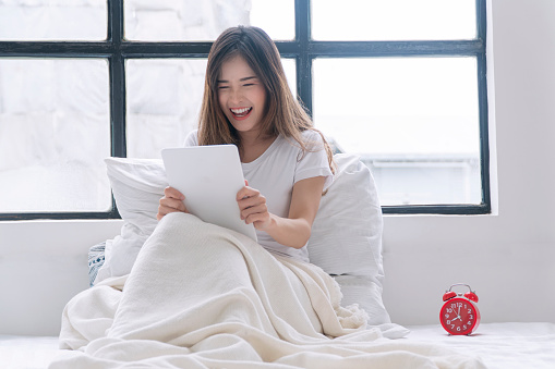 Excited asian woman play gaming online with tablet sitting beside window at home.Joyful young girl holding tablet touchpad playing game application fun relaxed with pillow indoors.Lifestyle technology