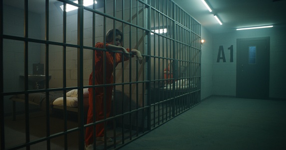 One female prisoner in orange uniform stands behind metal bars, another sits on the bed in prison cell. Women serve imprisonment terms for crimes in jail. Depressed inmates in correctional facility.