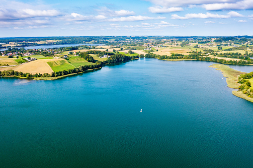 A large lake from above with a few small islands and a shoreline with trees and houses in Kashubia, Poland. The lake is a deep blue color and the sky is a bright blue with white clouds. The shoreline has a mix of trees and houses, and there are a few small boats on the water. The horizon is visible in the distance with a few more islands. The photo captures the beauty and tranquility of the natural landscape.