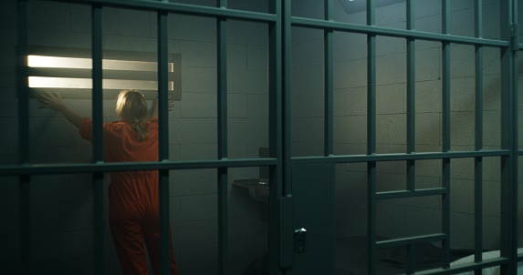 Female prisoner in orange uniform walks in jail cell, looks at barred window. Another woman leans on metal bars in neighbor prison cell. Women inmates in detention center or correctional facility.