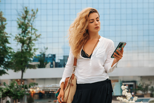 Woman with blond hair stands next to an office building and looking an at the phone in her hands