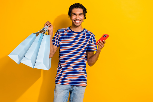 Portrait of toothy beaming man with piercing wear stylish t-shirt hold shopping bags smartphone isolated on yellow color background.