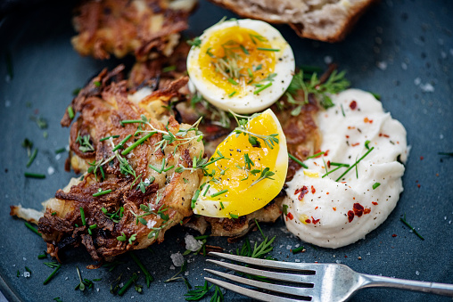 Bowl of freshly made latkes or potato cakes served with soft boiled eggs, 6 minutes, garlic mayonnaise and seasoned with salt and pepper, dill and chives and a slice of sourdough bread. The latkes are made with, grated potato, white onion, egg, flour and some creamed horseradish sauce. Then the individual cakes are shallow fried in sunflower oil. It is important to strain as much water out of the potatoes as possible. Colour, horizontal format with some copy space.