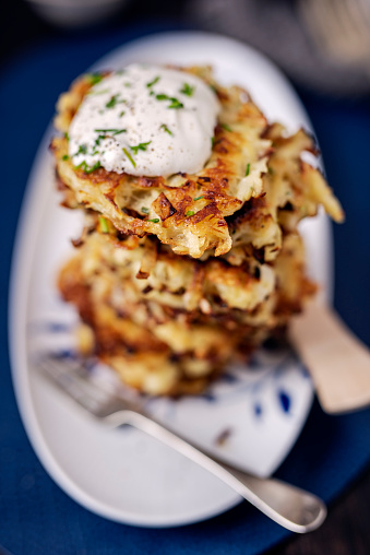 Pile of freshly made latkes or potato cakes. These are made with, grated potato, white onion, egg, flour and some creamed horseradish sauce. Then the individual cakes are shallow fried in sunflower oil. It is important to strain as much water out of the potatoes as possible. Here they are served with a sour cream dressing with fresh chives and dill. Colour, vertical format with some copy space.