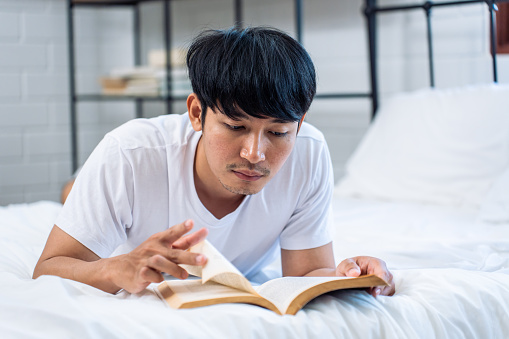 Asian handsome young man reading a book in the early morning at home. Happy attractive male lying down on bed in bedroom enjoy holiday weekend, using smartphone communication with technology in house.