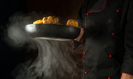 A professional chef prepares crispy nagits in a steamed hot pan. Recipe for delicious food and dishes on a black background. Molecular gastronomy or cuisine.