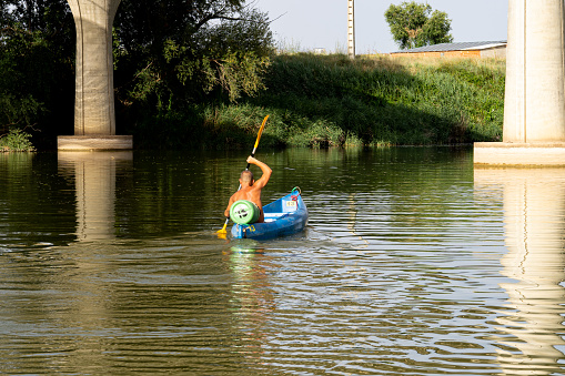 On a sunny summer afternoon, on the banks of the Duero River as it passes through Tordesillas-Spain and under a concrete bridge, a man paddles a blue canoe. Active life in nature. Nautical recreational activity.