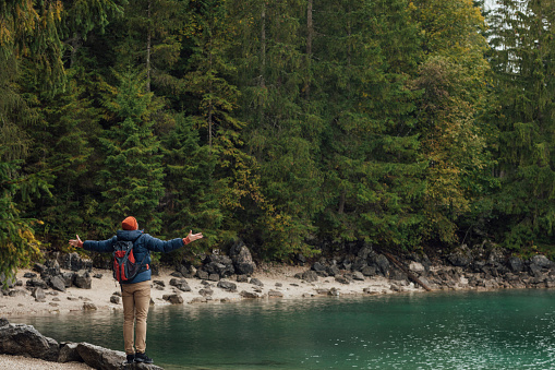A mid adult man wearing a waterproof and backpack exploring lake Eibsee while on holiday in Garmisch-Partenkirchen, Germany. He is standing on a rock at the lake's edge with his arms outstretched and enjoying the nature around him.