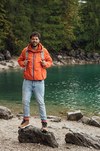 A portrait of a mid adult man wearing a waterproof and backpack exploring lake Eibsee while on holiday in Garmisch-Partenkirchen, Germany. He is standing on a rock with the lake behind him while he looks at the camera and smiles.