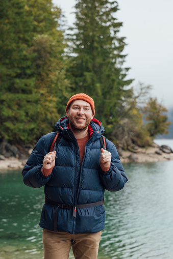 A portrait of a mid adult man wearing a padded jacket exploring lake Eibsee while on holiday in Garmisch-Partenkirchen, Germany. He is standing with the lake behind him while he looks at the camera and smiles.